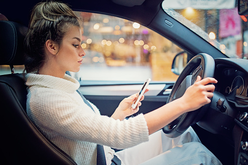 Young driver looking down at a cell phone while driving in the city. She's got one hand holding her phone and the other on the steering wheel of her car to indicate distracted driving.