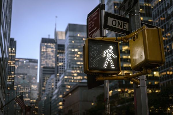 Illuminated pedestrian walk signal at dusk in a busy downtown area surrounded by tall glass buildings.