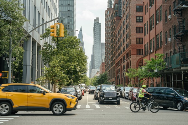 A busy street in New York City lined with tall buildings and trees, showing multiple lanes of traffic and a cyclist in the foreground riding across a crosswalk.