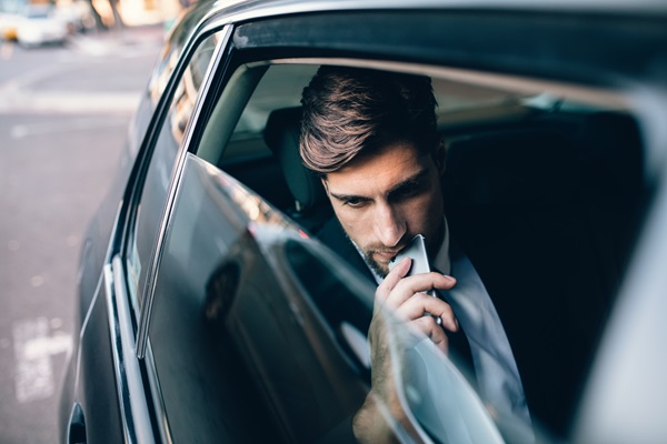 A man in a suit sitting in the back of an Uber vehicle, looking pensively out the window while holding a smartphone to his chin and being driven through New York City.