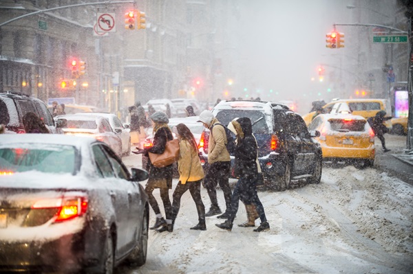 Pedestrians crossing a snowy New York City street filled with heavy traffic and yellow taxis during a winter storm.