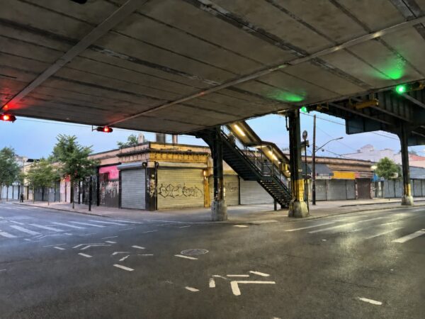 An empty street intersection beneath an elevated subway track line in the Bronx, featuring outdoor transit staircases and closed storefronts with security grates at dawn, representing the scene of a hit-and-run pedestrian accident and the legal options available for injured victims pursuing personal injury claims.